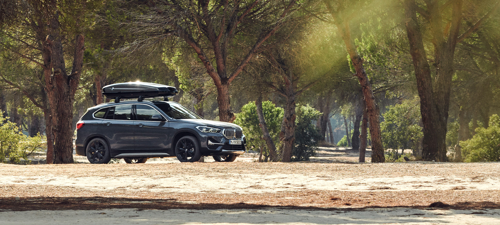 Side view BMW X1 with BMW Roof box BMW offers summer Side view BMW X1 Mineral Grey with BMW Roof box standing in forest BMW offers summer